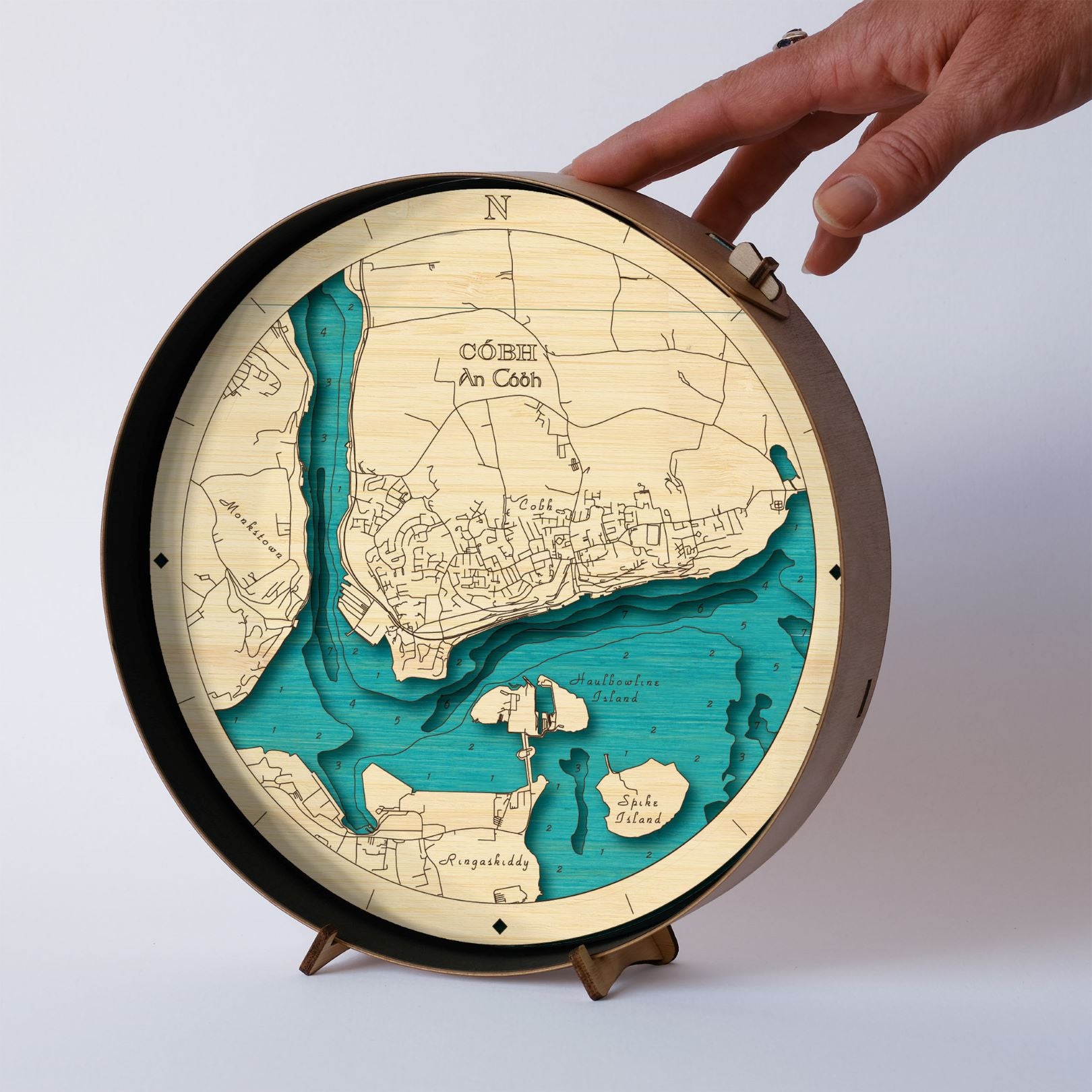 Round wooden map of Cobh on a wooden stand in a black frame with a neutral background and a hand touching the top