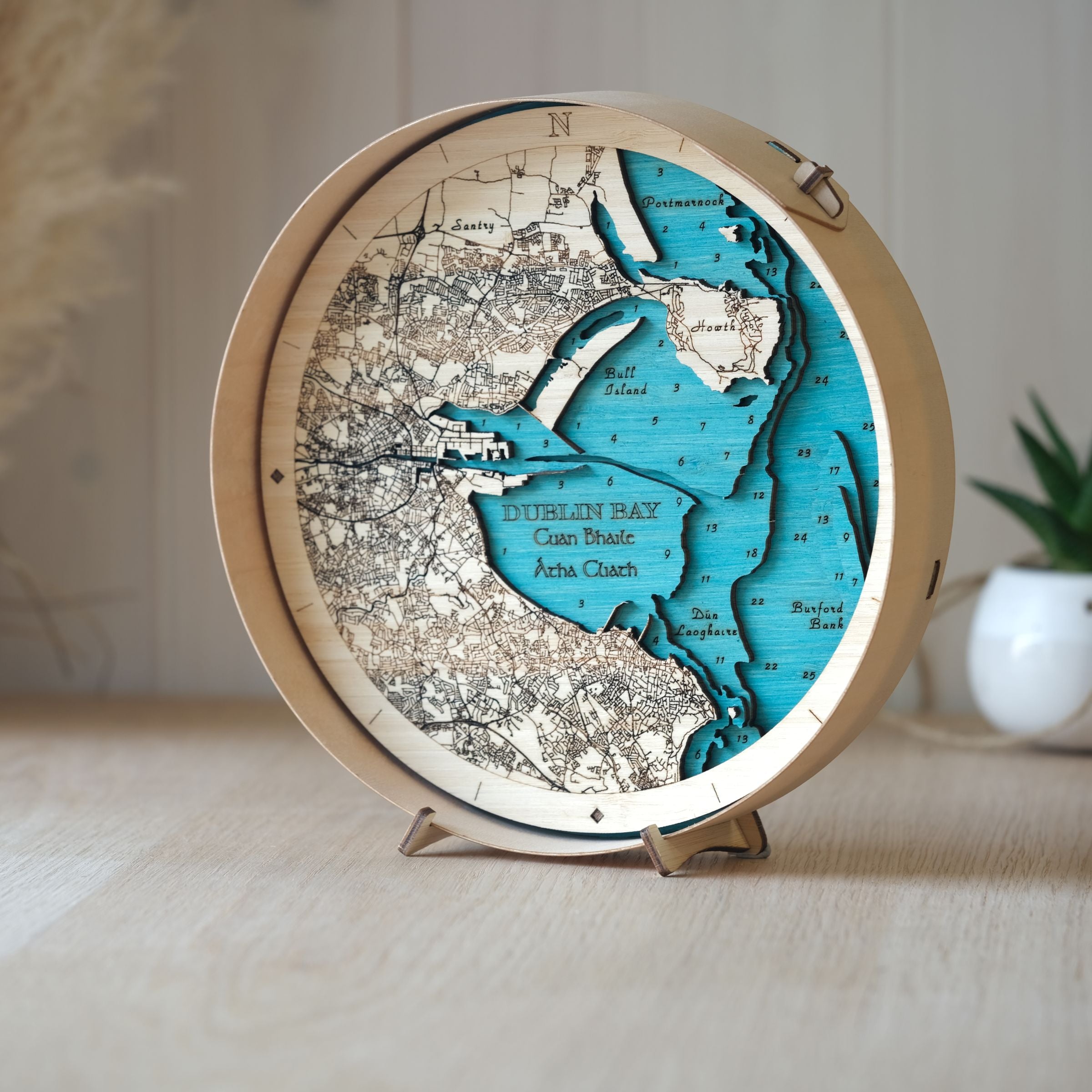 Handcrafted round wooden map of Dublin Bay in natural frame, displayed on an oak table with a decorative pot plant behind.