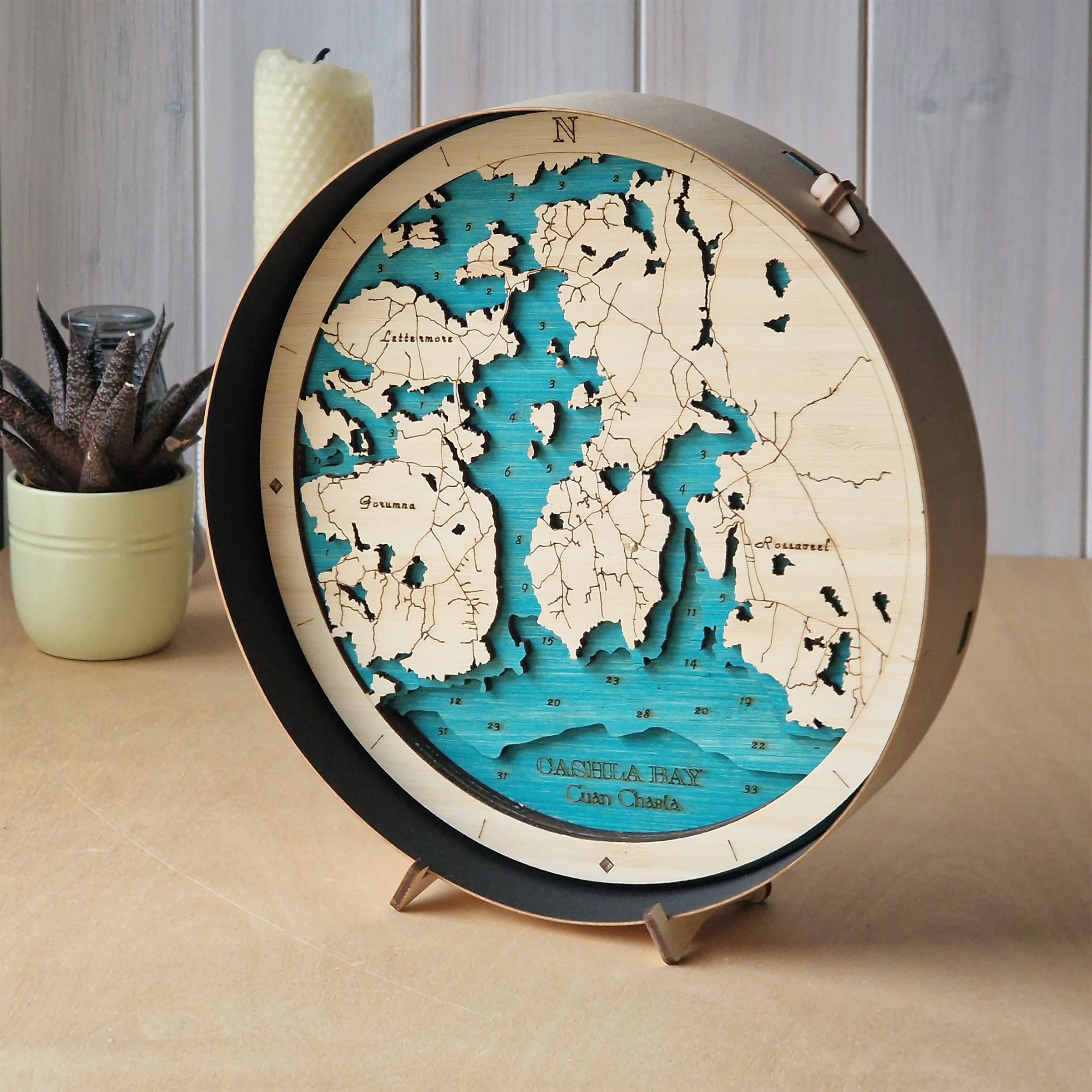 A round wooden 3D laser-cut map of Greece and surrounding islands, showcasing carved land layers in a light natural wood tone and sea layers in a vibrant turquoise blue. The map is displayed on a small metal stand on a beige tabletop, with a small potted succulent and a white candle in the background against a light wood paneled wall.