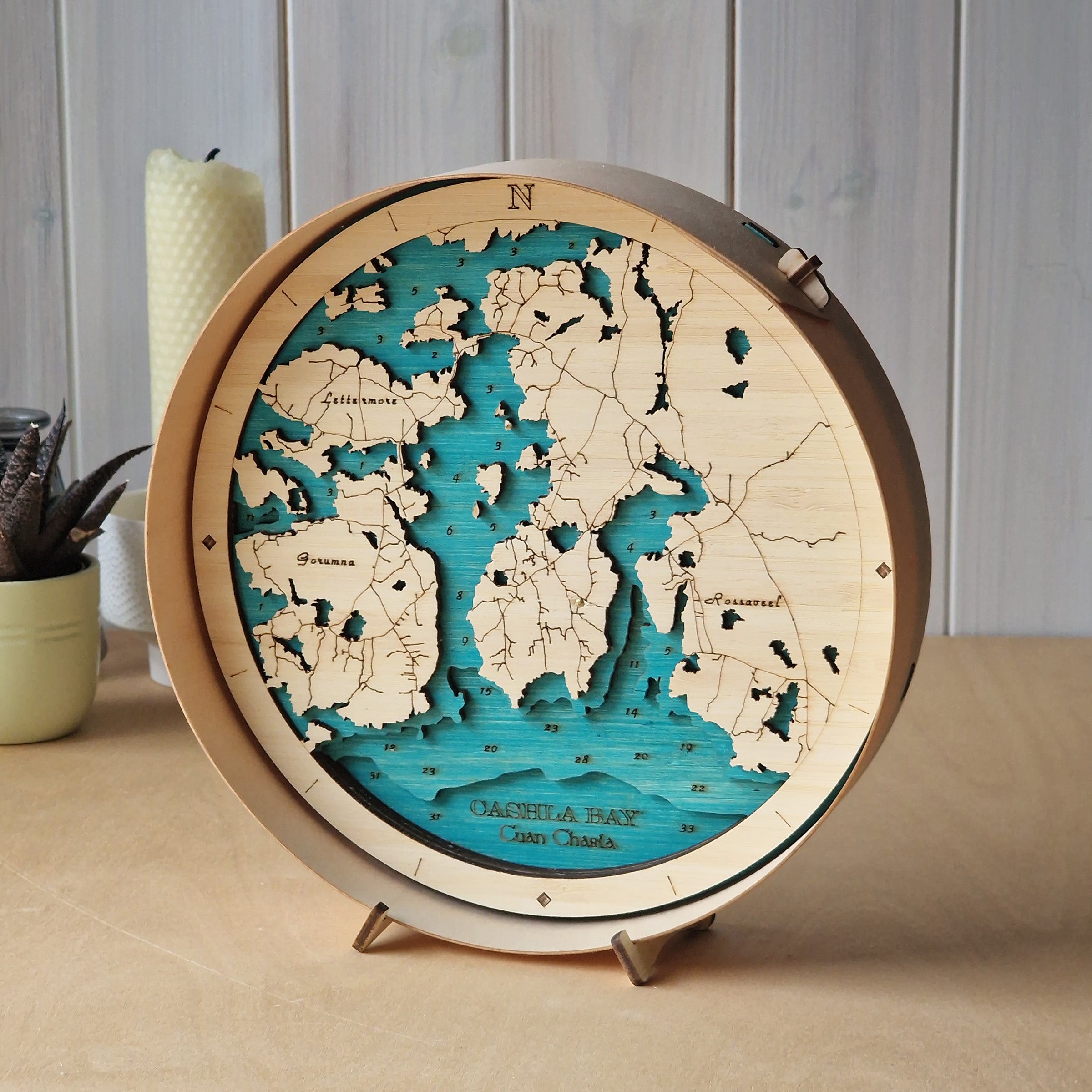 A round wooden world map stand with a 3D layered design showing continents in light natural wood and oceans in turquoise blue, displayed on a tabletop with a candle and potted plant in the background.
