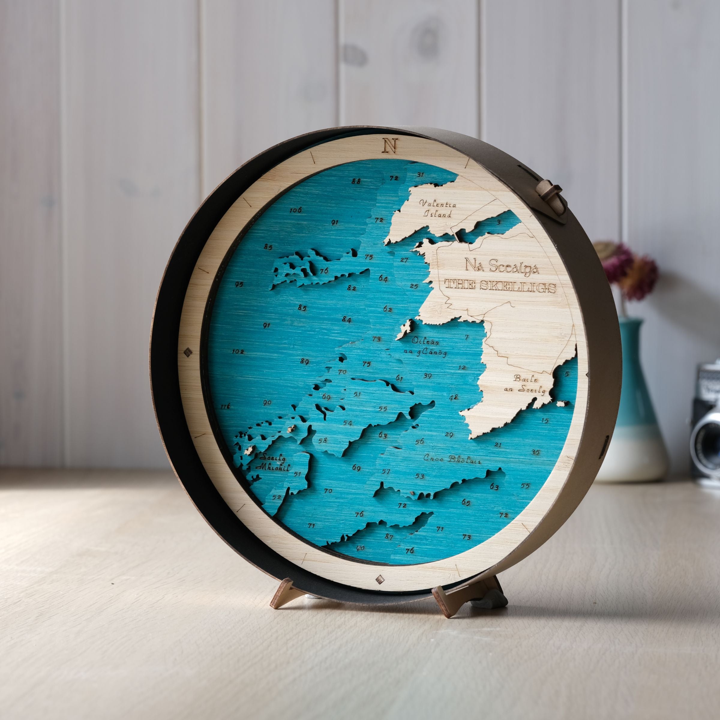 The Skelligs round wooden map in a black frame, showing Skellig Michael and Little Skellig, styled on a wall shelf in a bright, modern coastal interior.