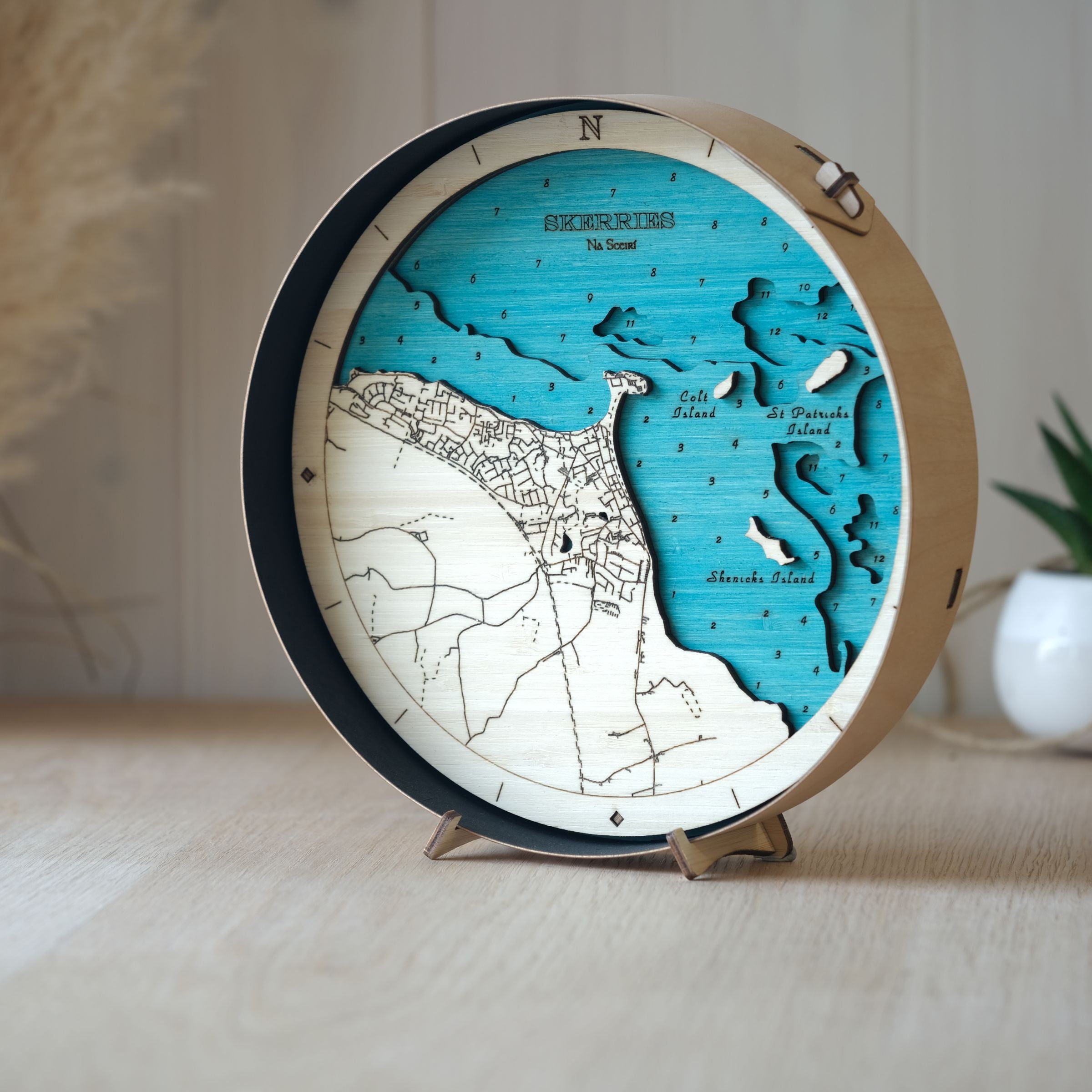 Round 3D wooden map of Skerries, framed in black wood, shown on an oak table with a small pot plant behind.