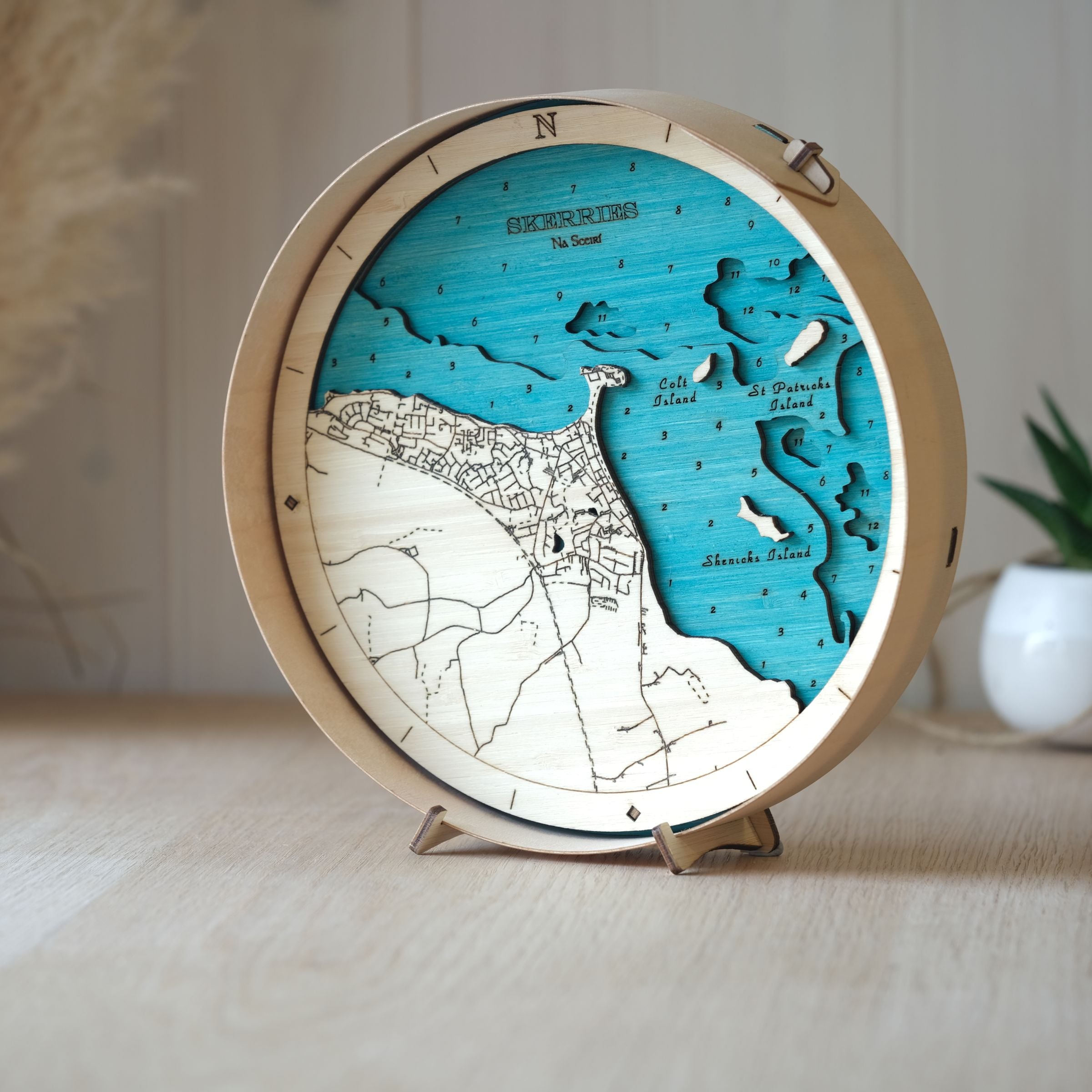Round 3D wooden map of Skerries, framed in natural wood, shown on an oak table with a small pot plant behind.