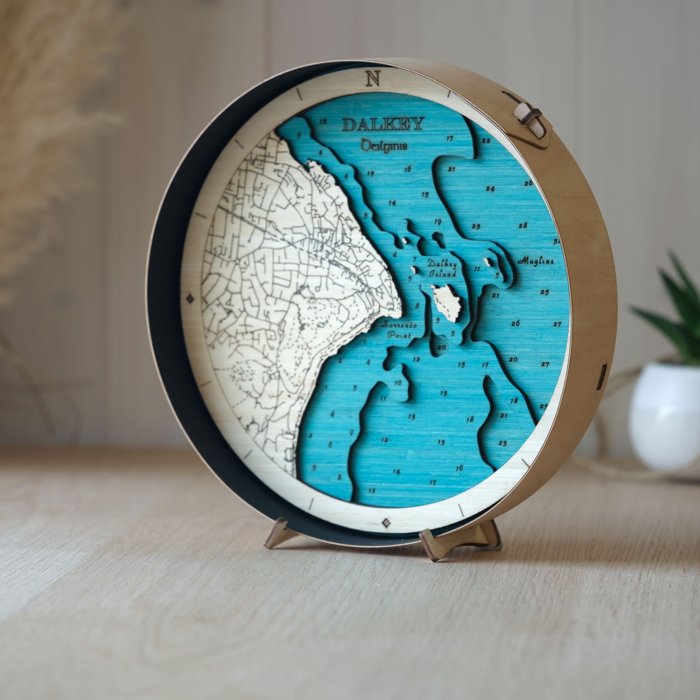 Handcrafted wooden map of Dalkey in a black wood frame, styled on an oak table with a pot plant behind for a cosy setting.