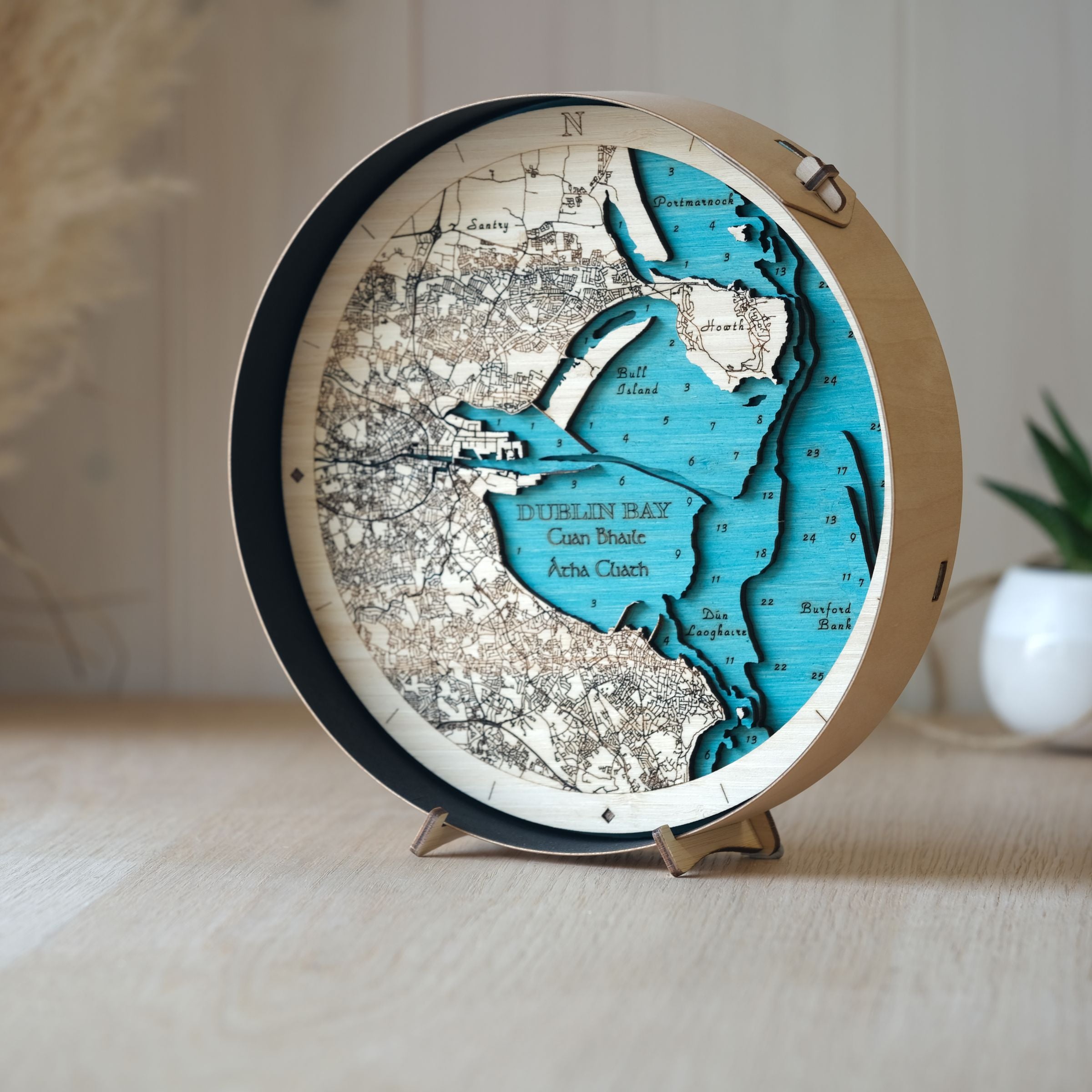 Handcrafted round wooden map of Dublin Bay in black frame, displayed on an oak table with a decorative pot plant behind.