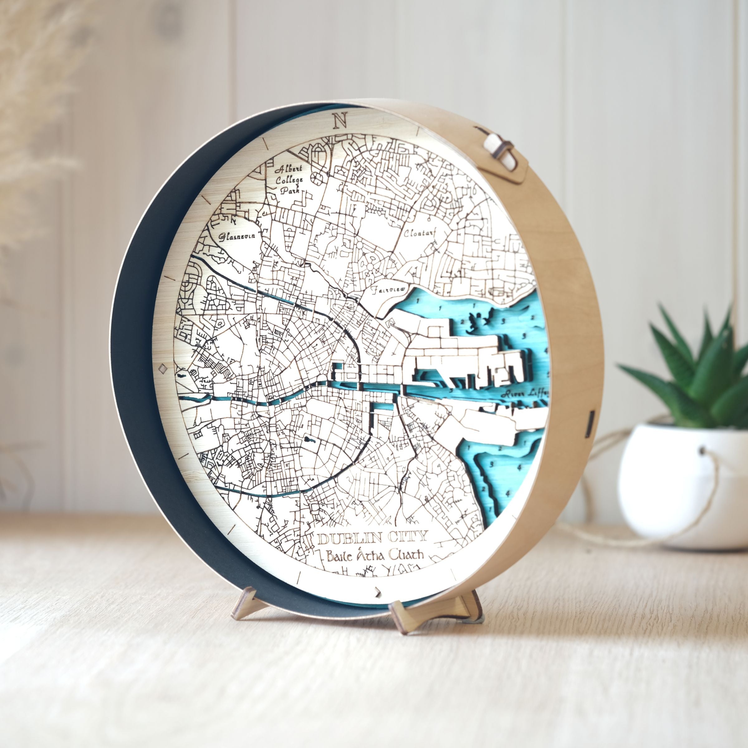 Round wooden map of Dublin City in a black wood frame, styled on an oak table with a pot plant in the background.