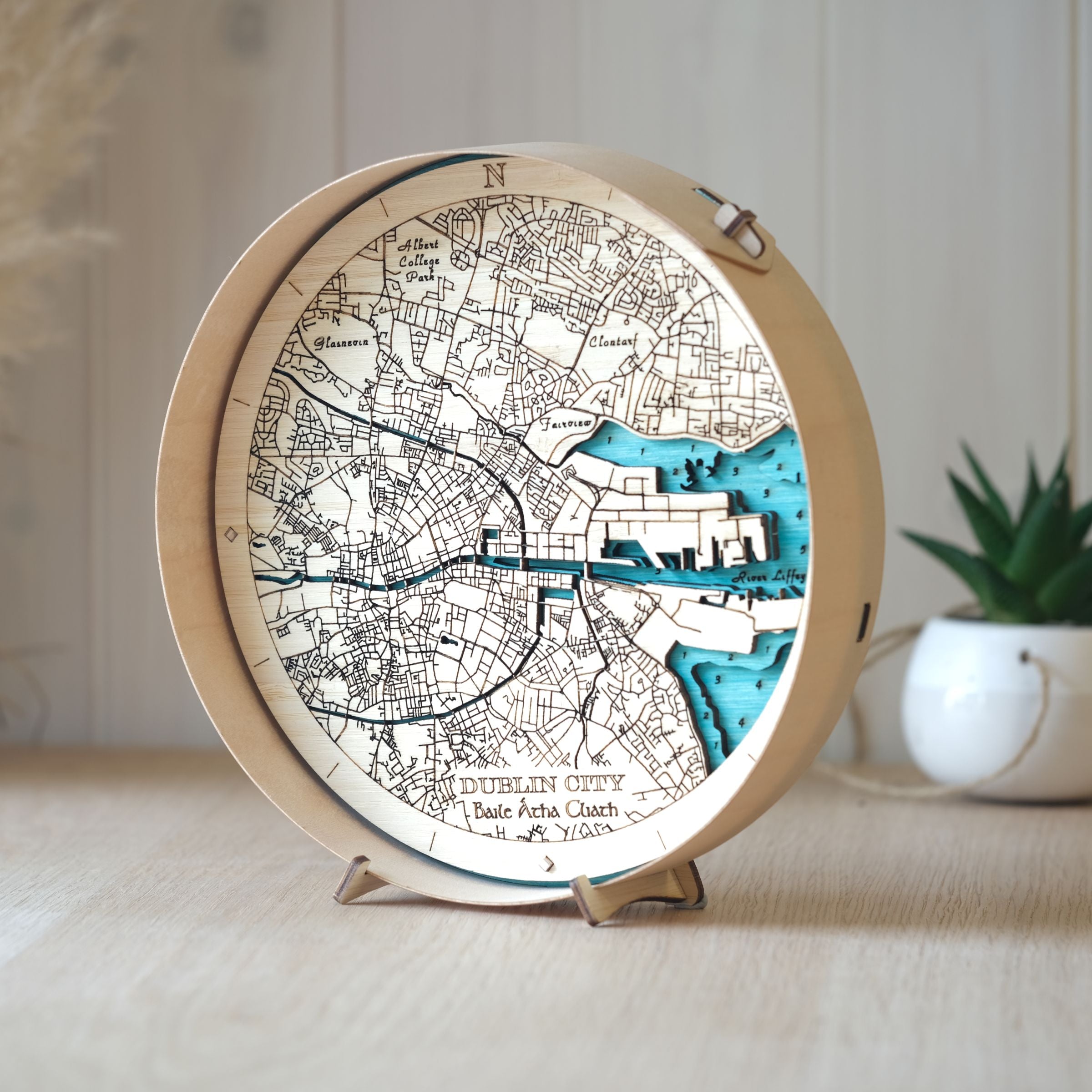 Round wooden map of Dublin City in a natural wood frame, styled on an oak table with a pot plant in the background.