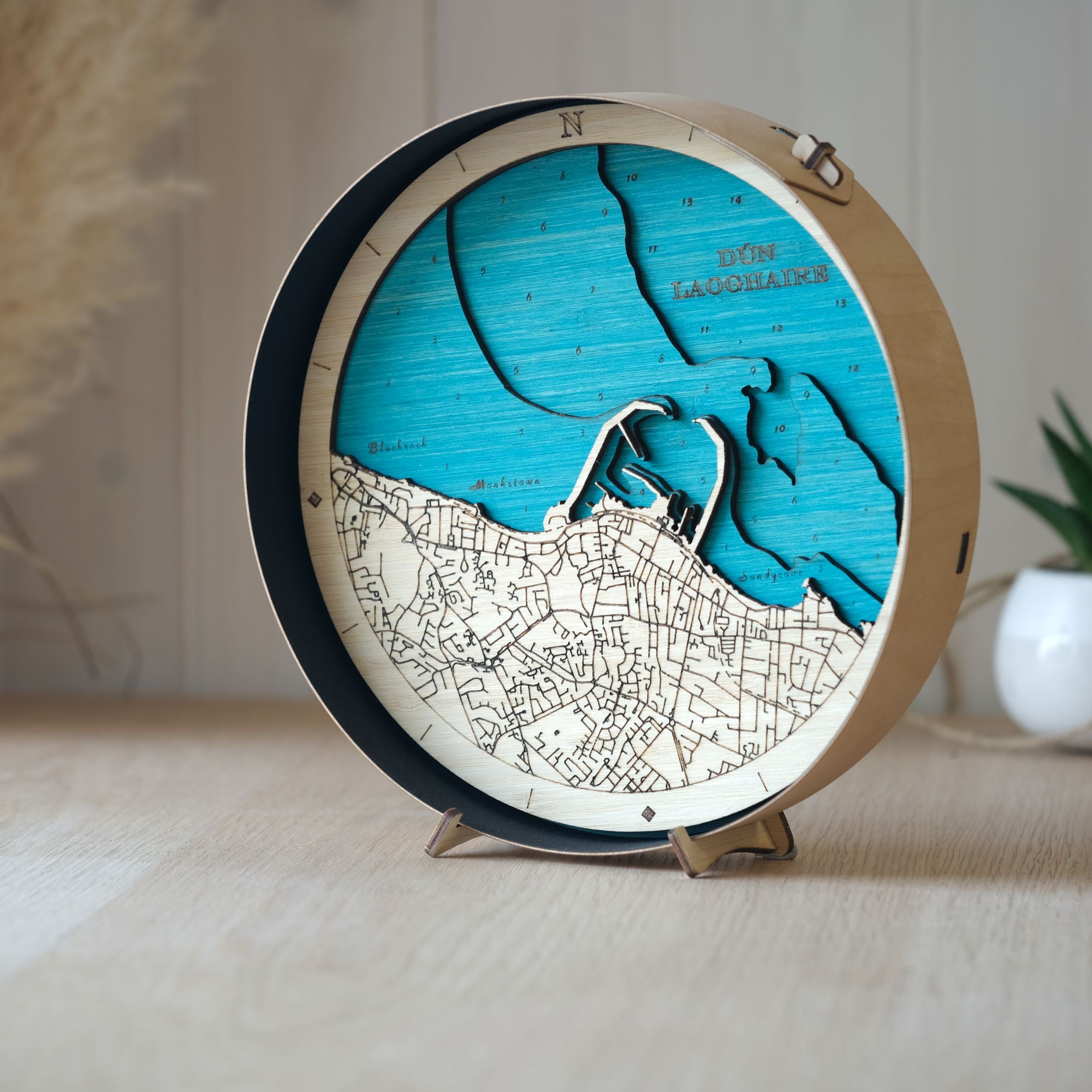 Round wooden map of Dún Laoghaire, black wood frame, styled on an oak table with a pot plant softly blurred in the background.