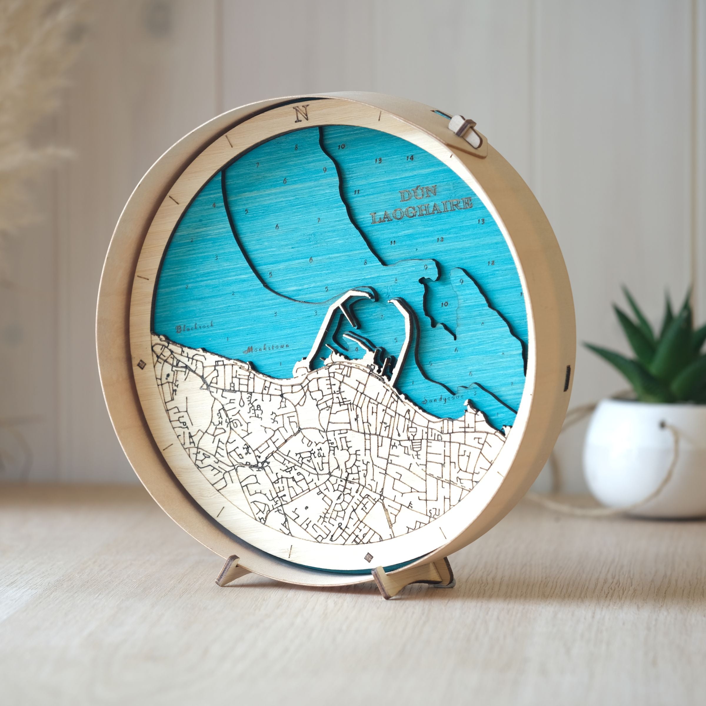 Round wooden map of Dún Laoghaire,  natural wood frame, styled on an oak table with a pot plant softly blurred in the background.