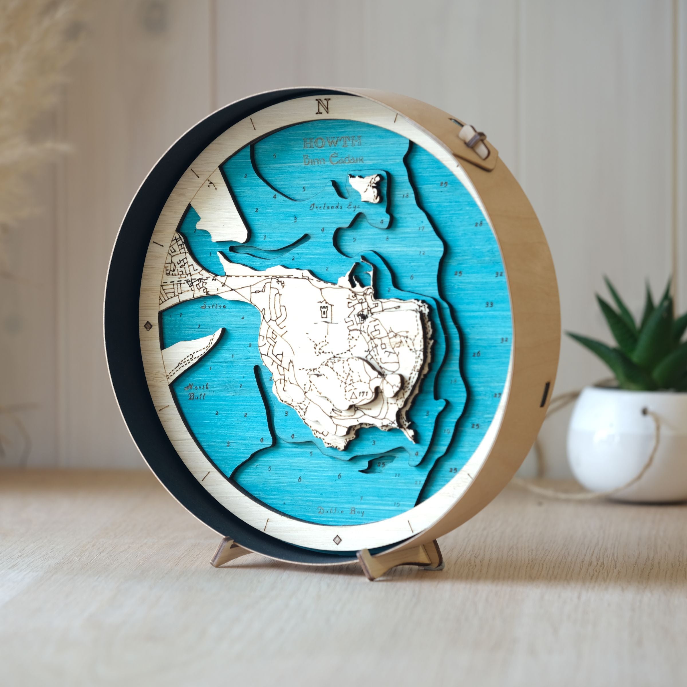 Beautifully layered wooden map of Howth, framed in black  wood, styled on an oak table with a pot plant in the background.