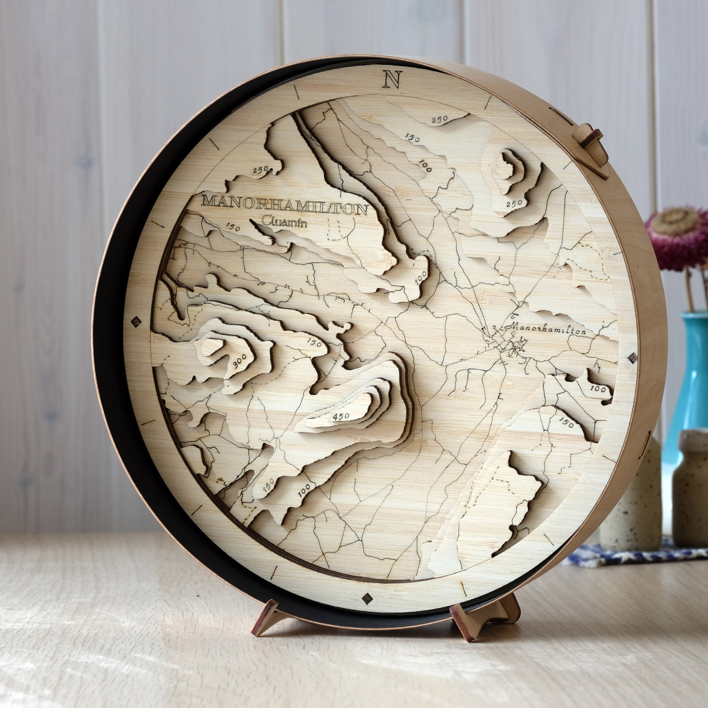 Manorhamilton round wooden map in a black frame, showing the town and surrounding Leitrim hills, styled on a wall shelf in a bright modern space.