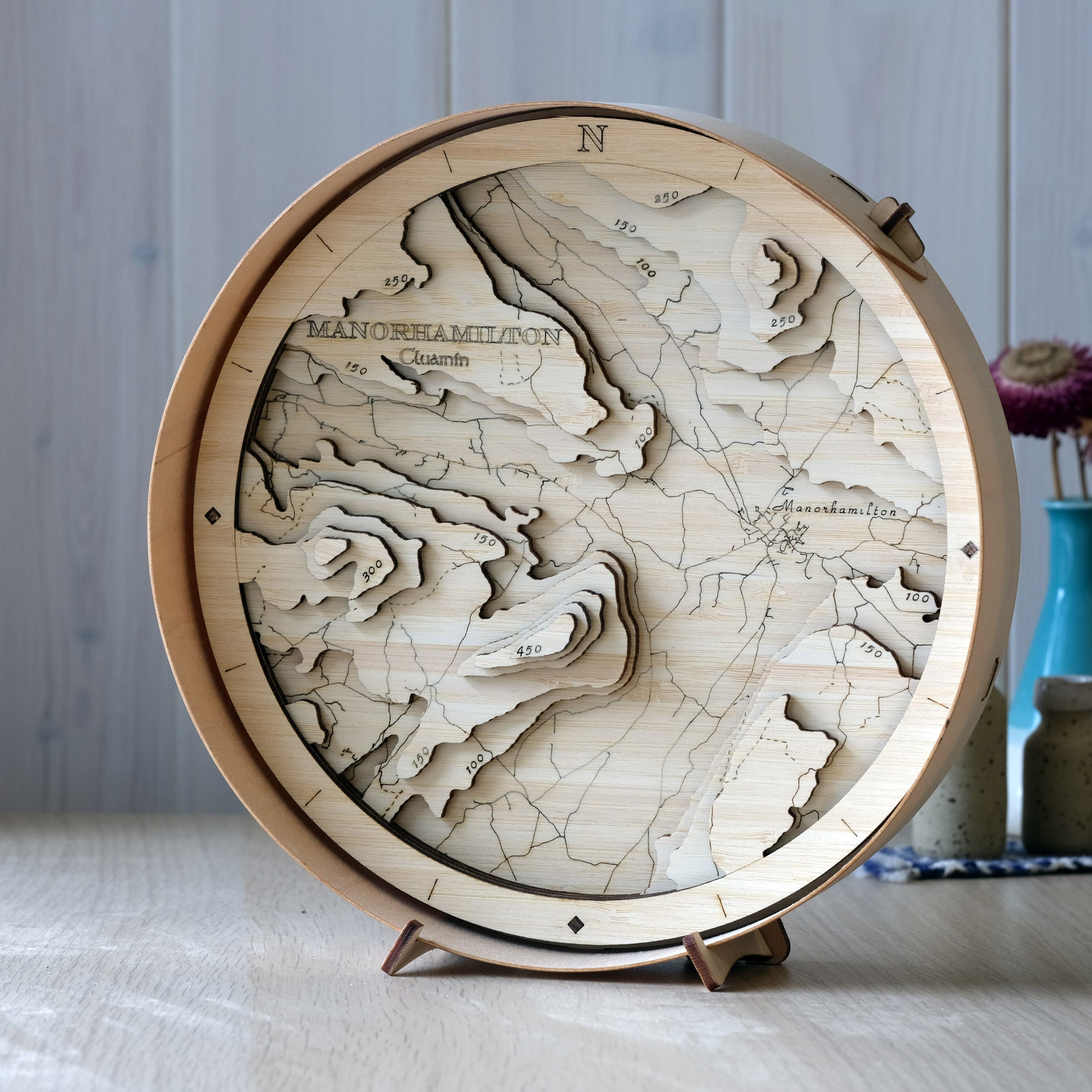 Manorhamilton round wooden map in a natural frame, showing the town and surrounding Leitrim hills, styled on a wall shelf in a bright modern space.