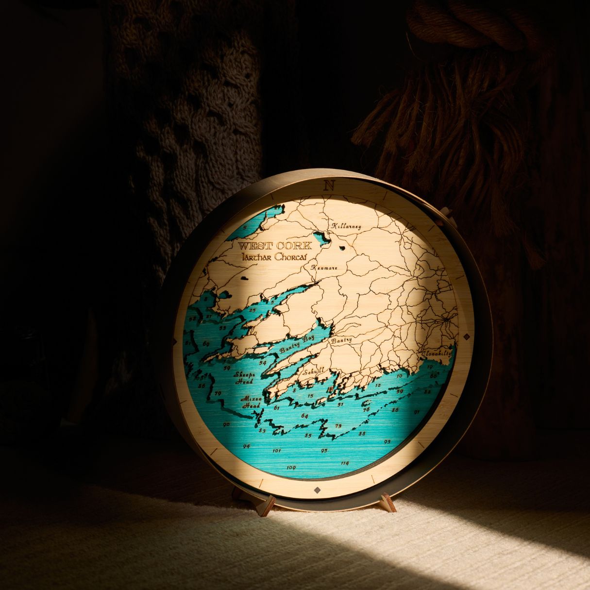 West Cork round wooden 3d map in a black frame, on a table with a sharp sliver of sunlight shining on it