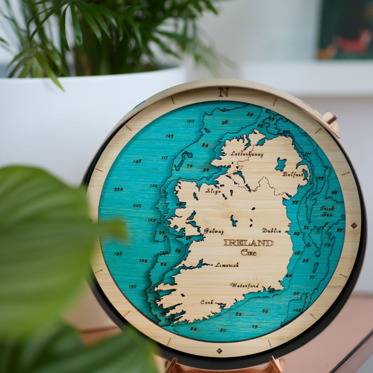 Round 3D wooden Ireland map displayed on a table with a green plant, showcasing sustainable Irish wall art in a home setting