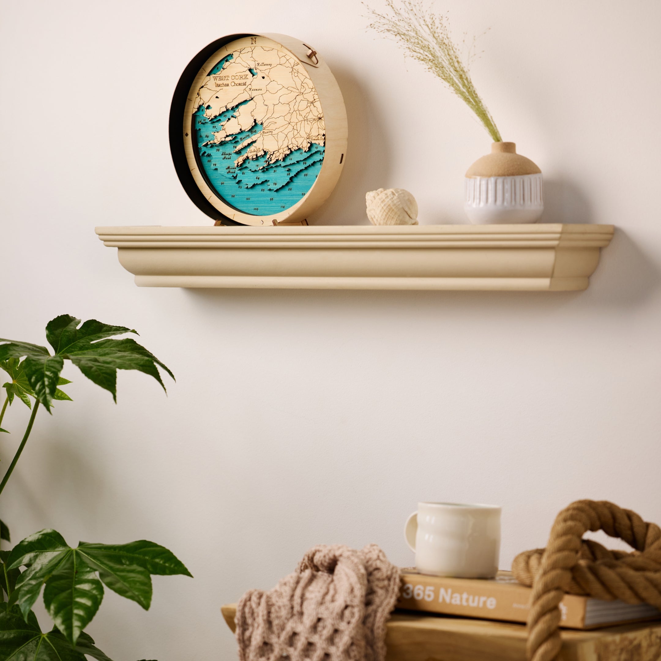 West Cork round wooden map in a  black frame, taken from a side angle styled on a wall shelf in a bright, modern sitting room with high-end minimalist decor.